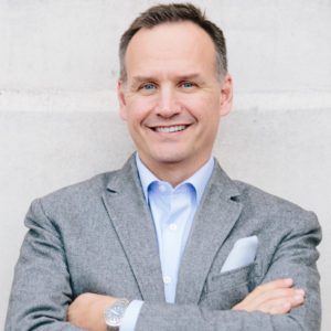 Matt Waters wearing grey blazer, blue shirt, pocket kerchief, no tie; smiling and looking into camera, arms crossed; standing against grey wall (color photo)