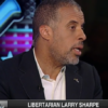 Larry Sharpe speaking, on the set of 'Kennedy', wearing dark sportcoat and white collared shirt, looking to his left, with large illuminated blue letter K for Kennedy in background, text on screen is 'Libertarian Larry Sharpe is running for Governor of New York' (color photo)