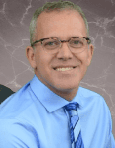 Rod Hanscomb portrait - wearing blue shirt and tie, eyeglasses, smiling, seated (color photo)